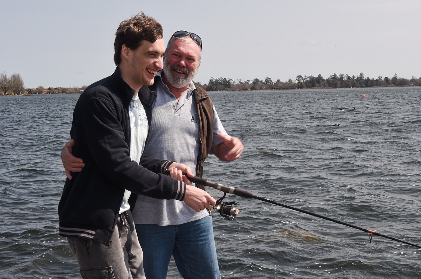 A support worker and a participant smiling while fishing together by a lake.