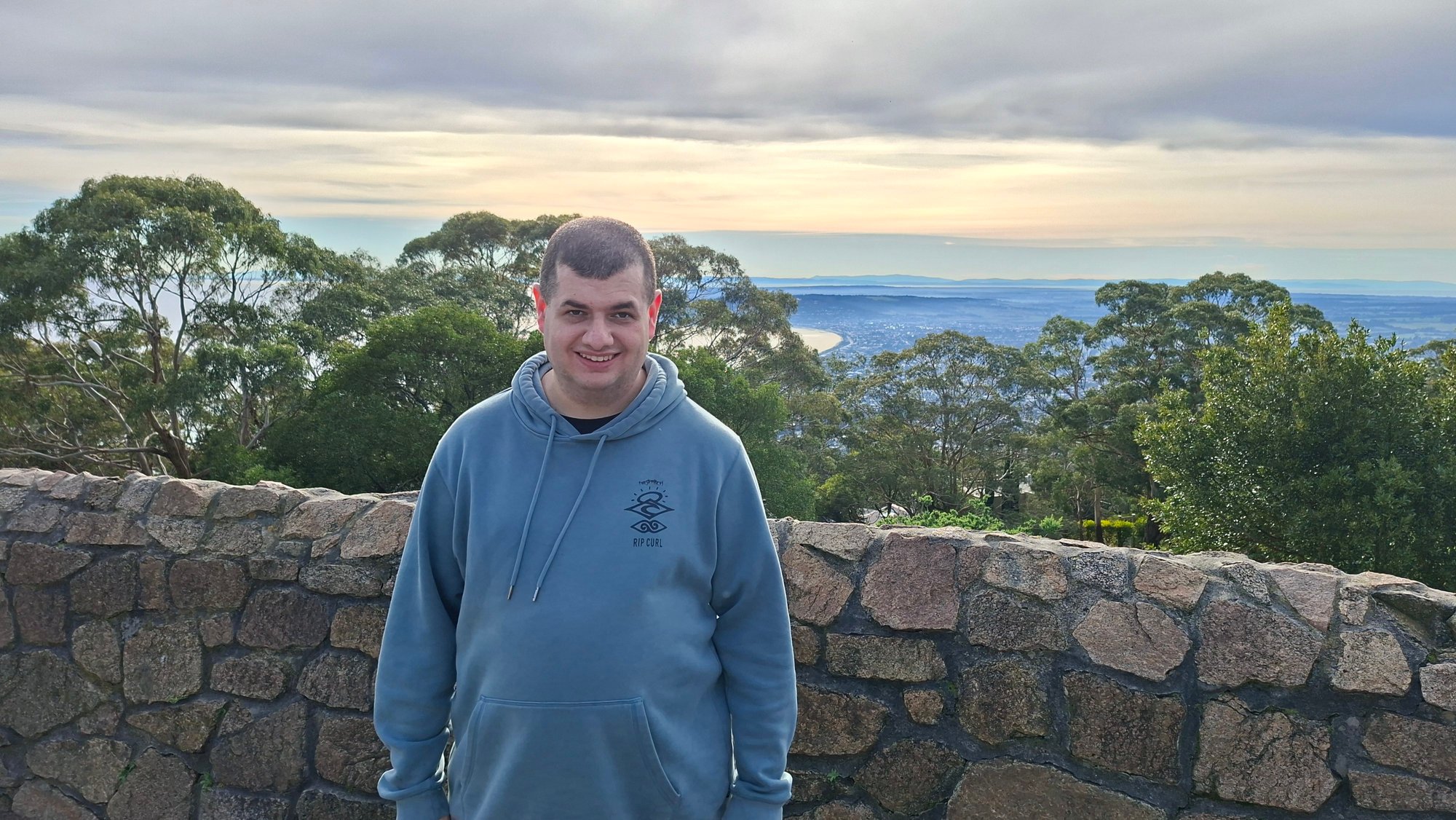 Man wearing a blue jumper in front of stone wall with trees and ocean in the background