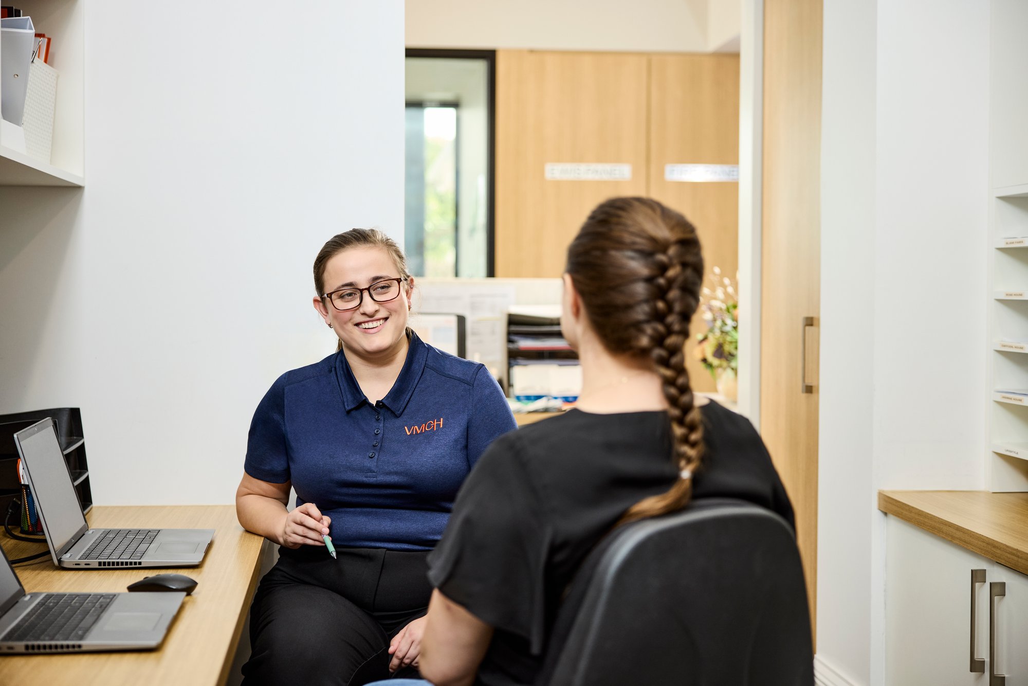 A woman talking to another at desk.