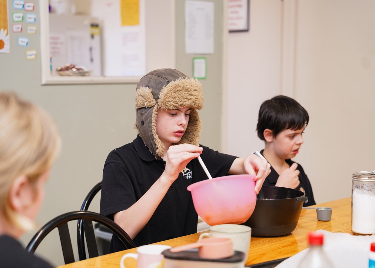 Two boys are sitting at the table. With cooking utensils in front of them, one boy is wearing a hat and is stirring with a wooden spoon in a pink mixing bowl.