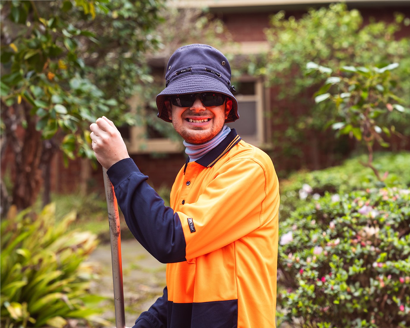 Man standing in garden holding rake handle wearing orange high vis work wear and navy hat