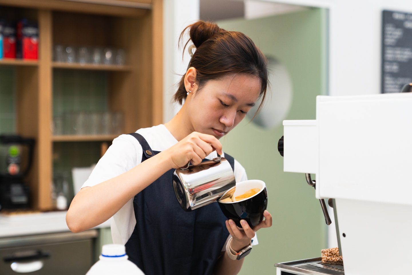 Female with dark hair wearing white tshirt and navy apron pouring milk into coffee cup