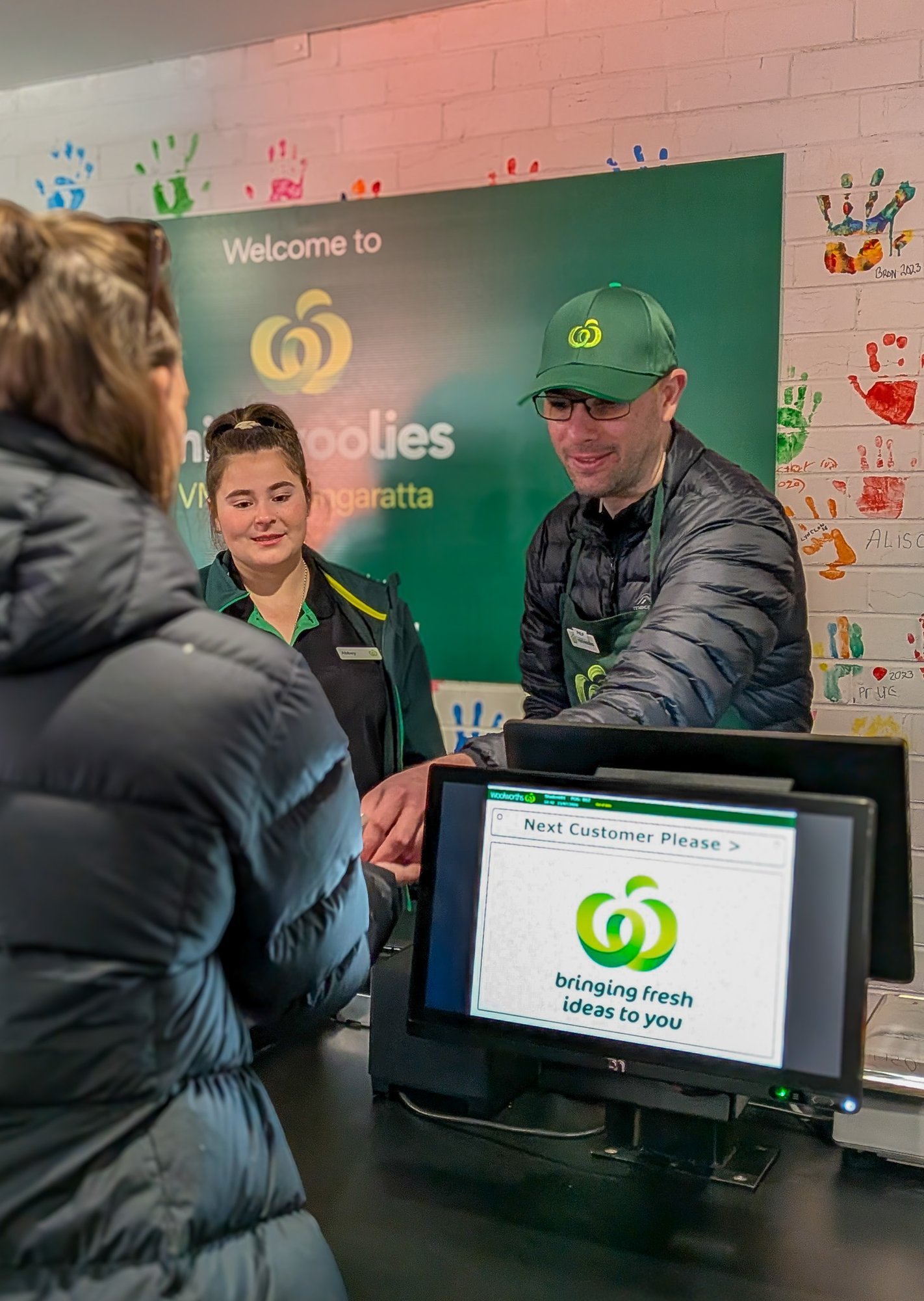 A man wearing a green cap and apron is standing behind a cash register, handing a person wearing a puffy jacket their change. Female with standing next to the man supervising