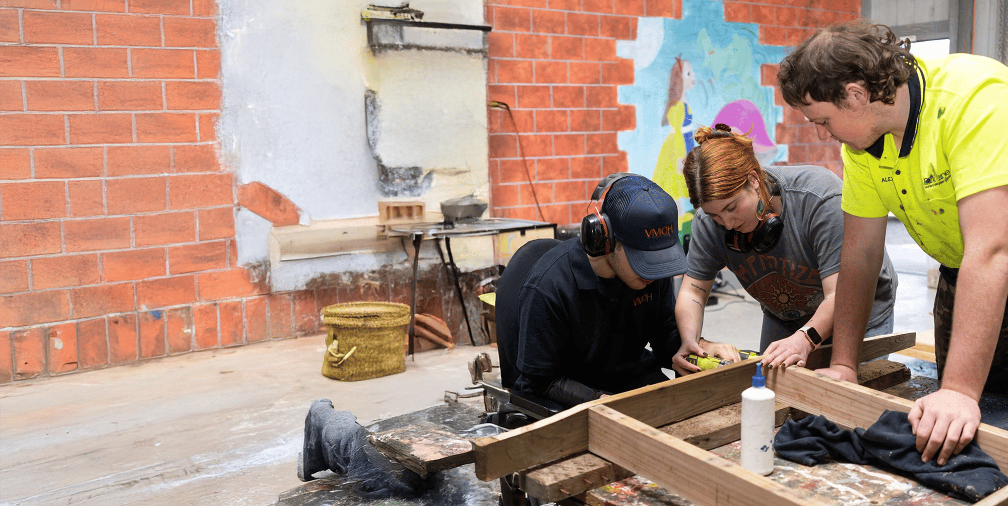 Three people working together on a woodworking project in a workshop, with tools and timber on the table, as part of a VMCH disability services program.