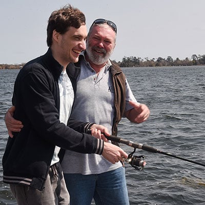 One man helping another hold a fishing rod by the ocean.